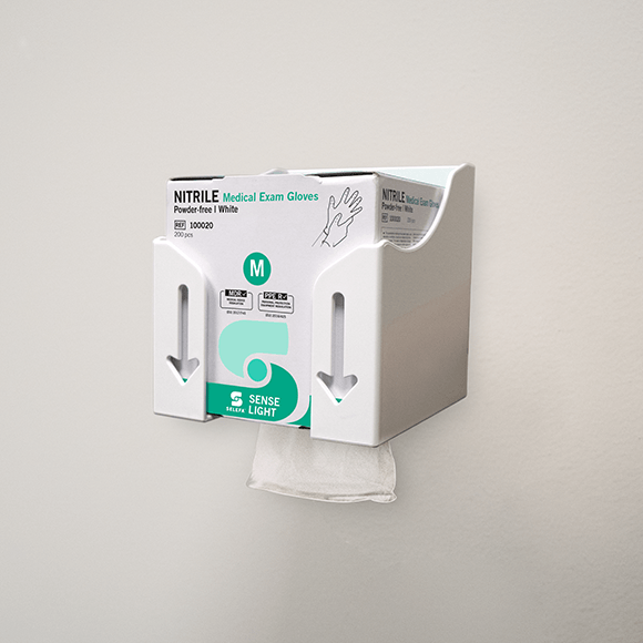 Wall-mounted white dispenser holding a box of medium-size powder-free white nitrile medical exam gloves, with one glove partially pulled out from the bottom.